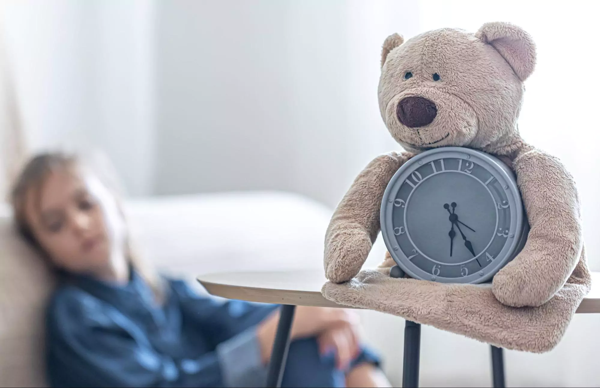 This image of a teddy bear with a clock and a child sleeping behind represents efficient time management for kids.